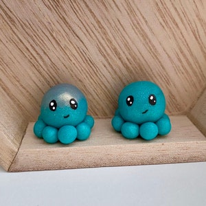 May include: Two small teal blue octopus figurines with white eyes and a sparkly finish. The octopus on the left has a silver shimmer on the top of its head.