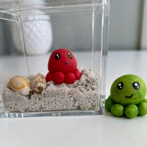 May include: A red octopus figurine inside a clear plastic container with sand and seashells. A green octopus figurine is next to the container. Both figurines have black eyes and smiling faces.