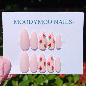 May include: Almond-shaped false nails in a pale pink colour, some decorated with a strawberry and daisy pattern. The card holding the nails displays the text "MOODYMOO NAILS".