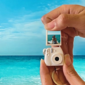 Puede incluir: Un llavero de cámara en miniatura blanco con una pequeña foto de una persona junto al mar. El llavero tiene un anillo plateado y se sostiene contra un fondo de cielo azul y océano. El texto "STL FILE" y "DIGITAL FILE" son visibles.