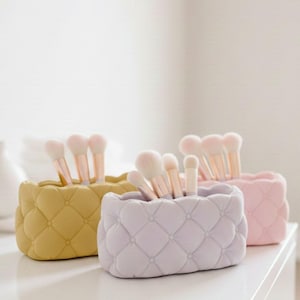 May include: Three rectangular makeup brush holders with a quilted, tufted texture, each featuring a series of small indentations. The holders are colored mustard yellow, light lavender, and soft pink, and contain various makeup brushes.