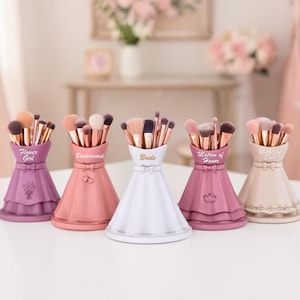 Può includere: Cinque porta pennelli per trucco a forma di abito, ciascuno pieno di pennelli. I supporti sono nei toni del viola, rosa e bianco. Il testo sui supporti recita "Flower Girl", "Bridesmaid", "Bride" e "Matron of Honor".