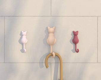 May include: Three cat-shaped hooks on a white wall. The hooks are pink, beige, and red. A brown wooden umbrella stand with a black umbrella is in the center.
