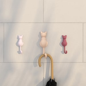 以下が含まれることがあります： 白い壁に猫の形をしたフックが3つ付いています。フックはピンク、ベージュ、赤です。中央には、黒い傘が付いた茶色の木製傘立てがあります。