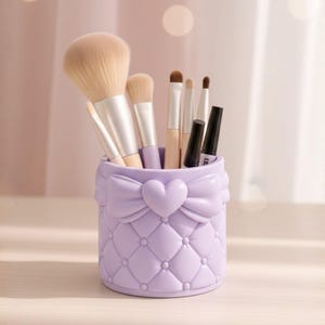 May include: A lavender makeup brush holder with a heart-shaped bow design. The holder is filled with various makeup brushes and black eyeliner. The text "HEART BOW MAKEUP HOLDER" and "3D STL FILE" are at the top.