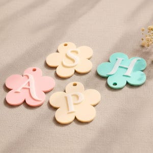 Peut inclure: Quatre breloques en forme de fleur aux couleurs pastel, chacune arborant une lettre blanche en relief. Les breloques comprennent une rose avec la lettre A, une jaune clair avec la lettre S, une jaune clair avec la lettre P et une bleu clair avec la lettre H.