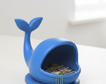 Puede incluir: Un organizador de joyas azul en forma de ballena con aleta caudal y base redonda. El vientre abierto de la ballena contiene varios anillos y collares. El texto "Whale Jewelry Organizer" y "3D PRINT FILES" está en la parte superior.
