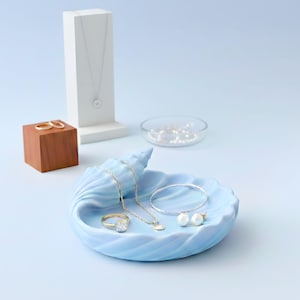 Könnte beinhalten: Hellblaue Muschelschalen-Schmuckablage mit Goldschmuck, darunter ein Ring, eine Halskette und ein Armband. Ein weißer Schmuckständer und ein kleiner Holzblock mit goldenen Ringen sind ebenfalls vorhanden. Der Text "CONCH SHELL JEWELRY TRAY" steht oben.