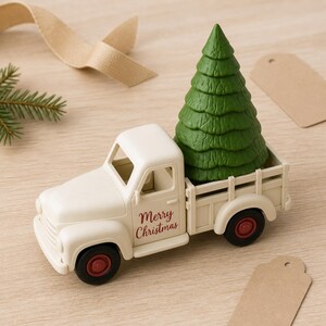Op de afbeelding: Een cr&egrave;mekleurige speelgoedpickup met de tekst Merry Christmas in het rood op de deur gedrukt. De laadbak van de truck vervoert een gestructureerde groene dennenboom. De wielen zijn zwart met rode centra en het voertuig heeft een klassiek ontwerp.