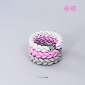 Op de afbeelding: Drie gevlochten armbanden gestapeld op elkaar. De bovenste armband is wit, de middelste armband is roze en de onderste armband is grijs.