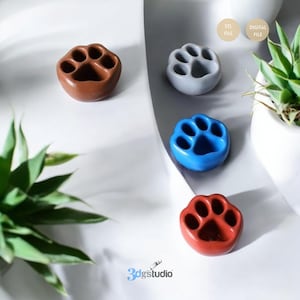 Peut inclure: Plusieurs porte-objets en céramique en forme de patte, de couleurs marron, grise, bleue et rouge. Ils sont disposés sur une surface blanche, avec une plante en arrière-plan. Chaque objet est en forme d'empreinte de patte.