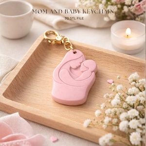May include: A pink keychain featuring a mother and child design, with the words "MOM AND BABY KEYCHAIN" and "3D STL FILE" printed above. The keychain is attached to a gold-colored clasp. The image is set on a wooden tray with flowers and a candle.
