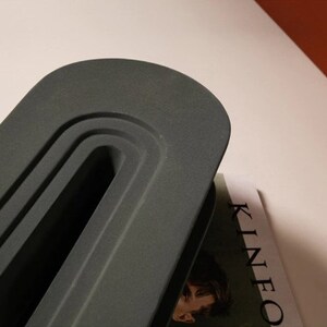 May include: A dark gray, oval-shaped object with a recessed, curved design. The object is positioned over a magazine with the word "KINEO" visible. The background is a light color.