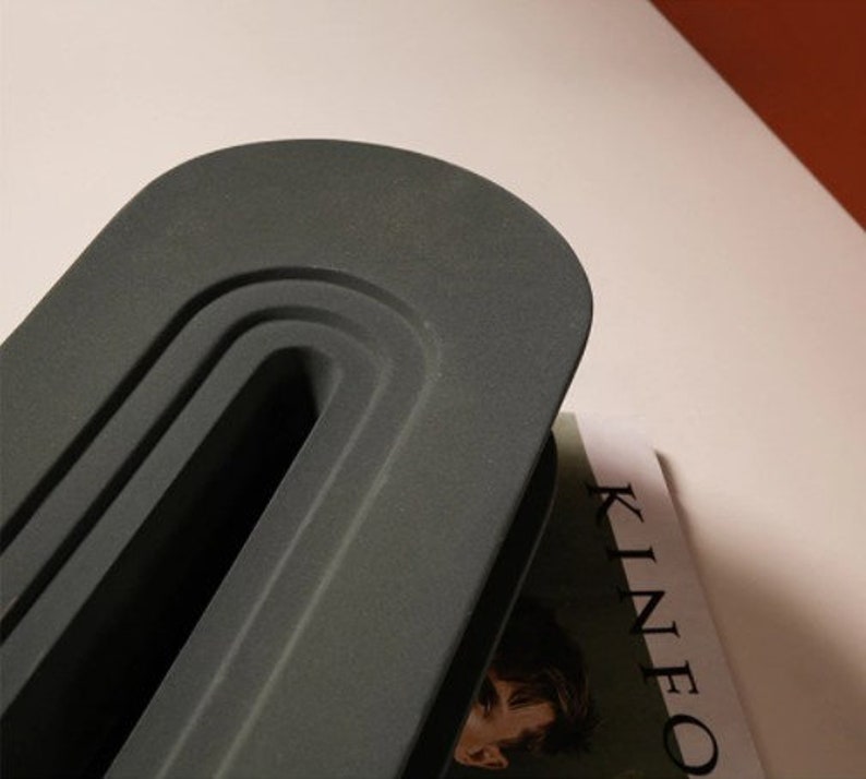 May include: A dark gray, oval-shaped object with a recessed, curved design. The object is positioned over a magazine with the word "KINEO" visible. The background is a light color.