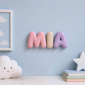 May include: A wall-mounted decorative sign spelling the name MIA in three-dimensional, rounded, bubble-style letters. The letter M is pink, the letter I is cream, and the letter A is light purple, all featuring a smooth, matte finish.