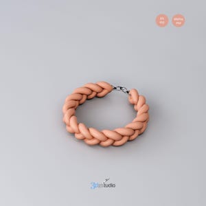 Op de afbeelding: Een gewoven armband in perzikkleur met een zilveren sluiting. De armband is gemaakt van een dik, touwachtig materiaal en is ontworpen om om de pols te dragen.