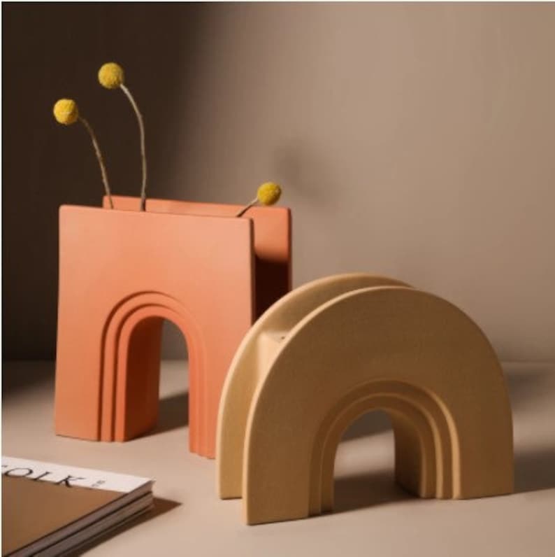 May include: Two ceramic vases in a modern design. One is a peach color, the other is a light yellow. The peach vase has a rectangular shape with an arched cutout. The yellow vase has a semi-circular shape with an arched cutout. The peach vase holds dried yellow flowers.