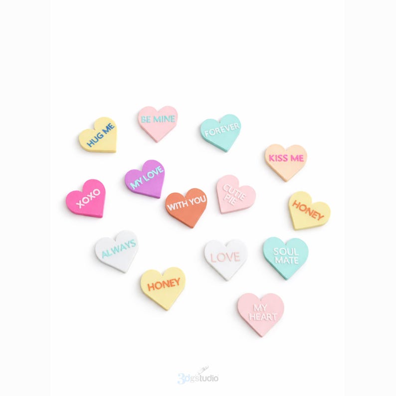 May include: A selection of heart-shaped sweets in pastel colours, each bearing a romantic message such as "HUG ME" and "BE MINE". The candies are scattered on a white surface, perfect for Valentine's Day or a romantic occasion.