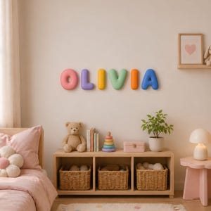 May include: A set of six individual, rounded, three-dimensional wall letters spelling the name OLIVIA in pastel colours. The letters are pink, purple, yellow, green, orange, and blue, mounted horizontally on a plain, light-coloured wall.