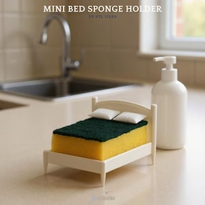 May include: A miniature white bed-shaped sponge holder with a yellow sponge and green scrub top. Two small white pillows rest on the sponge. A white soap dispenser is to the right. Text at the top reads "MINI BED SPONGE HOLDER" and "3D STL FILES".