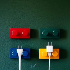 Peut inclure: Quatre supports muraux colorés pour stations de recharge, rouges, verts, bleus et jaunes, ressemblant à de grosses briques de jouets. Deux ont des prises de charge insérées, avec des cordons blancs qui s'étendent vers le bas. Le fond est vert foncé uni.