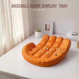 Peut inclure: Un plateau &agrave; bijoux en forme de gant de baseball orange. Le plateau est fait d'un mat&eacute;riau souple et pr&eacute;sente un design matelass&eacute;. Plusieurs colliers, bagues et boucles d'oreilles sont expos&eacute;s sur le plateau. Le texte "BASEBALL GLOVE JEWELRY TRAY" est visible.