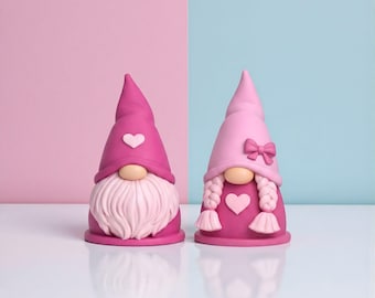 May include: Two Valentine's Day gnome figurines. One gnome has a pink hat with a white heart and a white beard. The other gnome has a light pink hat with a bow and braids. Both gnomes have a heart on their chest. The background is split between pink and blue.