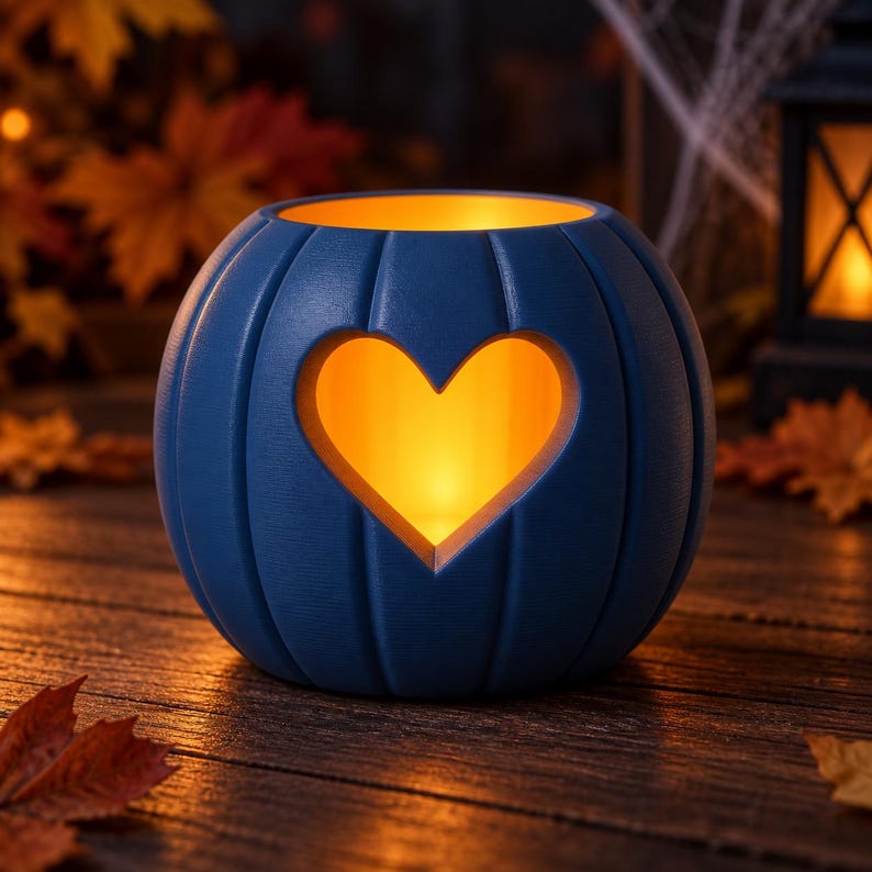May include: A round, blue, pumpkin-shaped candle holder with vertical ridges and a heart-shaped cutout in the center. The interior glows with a warm, golden light, illuminating the heart opening and the top rim of the blue pumpkin vessel.