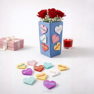 May include: A blue vase filled with red roses and heart-shaped decorations bearing phrases such as "Be Mine" and "Kiss Me." Additional heart decorations with phrases like "My Love" and "Forever" are scattered around. A pink gift box and a red candle are also visible.