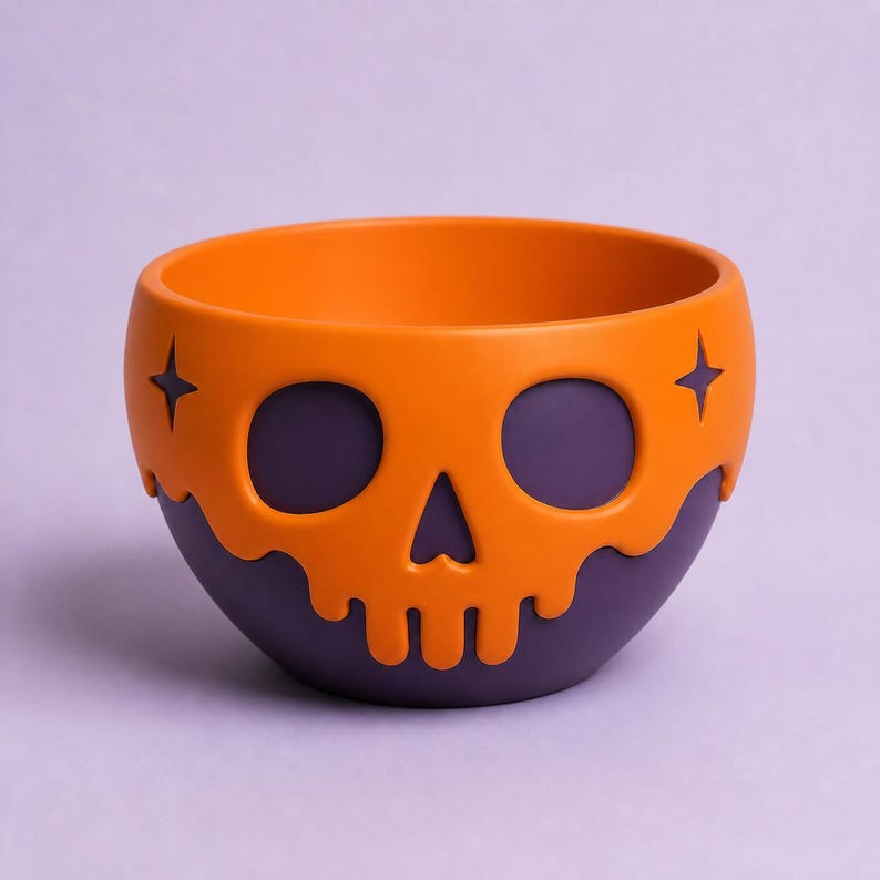 May include: A round bowl featuring a purple base and an orange overlay shaped like a skull with two four-pointed star accents. The orange skull design has a dripping effect along the top edge, creating a contrast against the solid purple bowl.