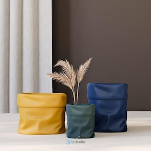 Può includere: Tre vasi in tessuto di diversi colori e dimensioni. Il vaso più grande è blu, il vaso centrale è verde e il vaso più piccolo è giallo. Una pianta secca è nel vaso centrale.
