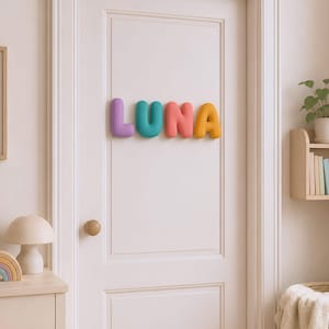 May include: A set of four individual, three-dimensional, rounded letters spelling the name LUNA, mounted on a white door. The letter L is purple, the U is teal, the N is coral pink, and the A is yellow. Each letter has a smooth, matte finish.