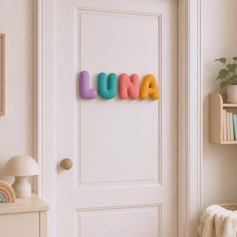 May include: A set of four individual, three-dimensional, rounded letters spelling the name LUNA, mounted on a white door. The letter L is purple, the U is teal, the N is coral pink, and the A is yellow. Each letter has a smooth, matte finish.