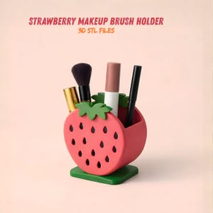 May include: A red strawberry-shaped makeup brush holder with green leaves and a green base. The holder contains various makeup brushes and tubes. Text at the top reads "STRAWBERRY MAKEUP BRUSH HOLDER" and "3D STL FILES".