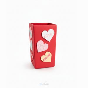 May include: A red rectangular container decorated with heart-shaped motifs. The hearts display phrases such as "LOVE", "CUTIE PIE", and "KISS ME". The container is a solid red colour with a smooth finish, set against a white backdrop.