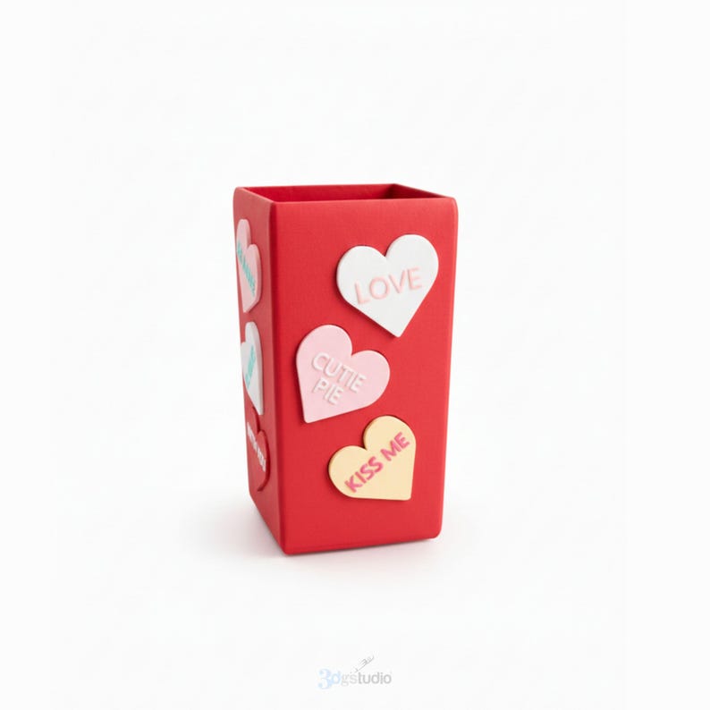 May include: A red rectangular container decorated with heart-shaped motifs. The hearts display phrases such as "LOVE", "CUTIE PIE", and "KISS ME". The container is a solid red colour with a smooth finish, set against a white backdrop.