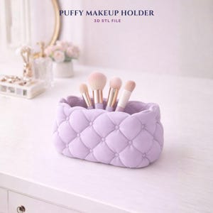Könnte beinhalten: Ein lavendelfarbener, gesteppter Make-up-Pinselhalter mit einem puffigen Design, der mehrere Make-up-Pinsel enthält. Der Text "PUFFY MAKEUP HOLDER 3D STL FILE" befindet sich oben im Bild. Der Halter steht auf einer weißen Oberfläche.