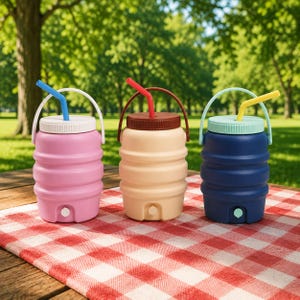 Op de afbeelding: Drie kleurrijke, opvouwbare waterflessen met rietjes en handvatten liggen op een rood-wit geblokt picknickkleed. De flessen zijn roze, beige en blauw, met bijpassende deksels en rietjes. De achtergrond is een groen park.