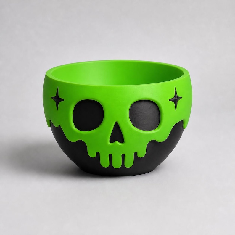May include: A round bowl featuring a matte black base and a bright lime green upper section. The green portion is shaped like a skull with black eye sockets and a nose cavity, accented by two small black four-pointed stars on the sides.
