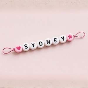 Puede incluir: Un clip para chupete con cuentas rosas y blancas con el nombre "SYDNEY" escrito en letras negras. El clip presenta una cuenta rosa en forma de coraz&oacute;n y estrella, y un lazo de alambre rosa.
