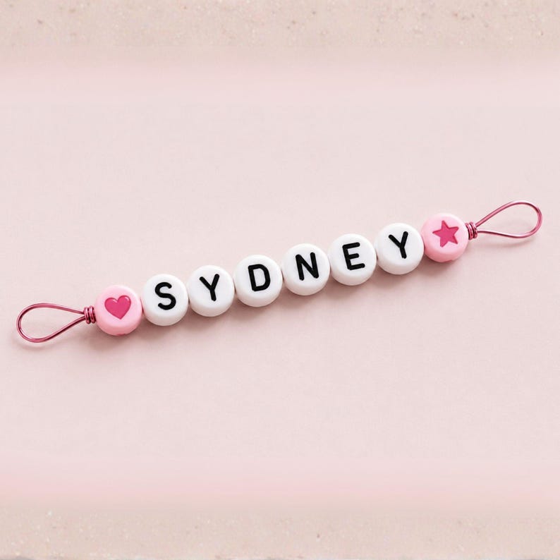 Puede incluir: Un clip para chupete con cuentas rosas y blancas con el nombre "SYDNEY" escrito en letras negras. El clip presenta una cuenta rosa en forma de coraz&oacute;n y estrella, y un lazo de alambre rosa.