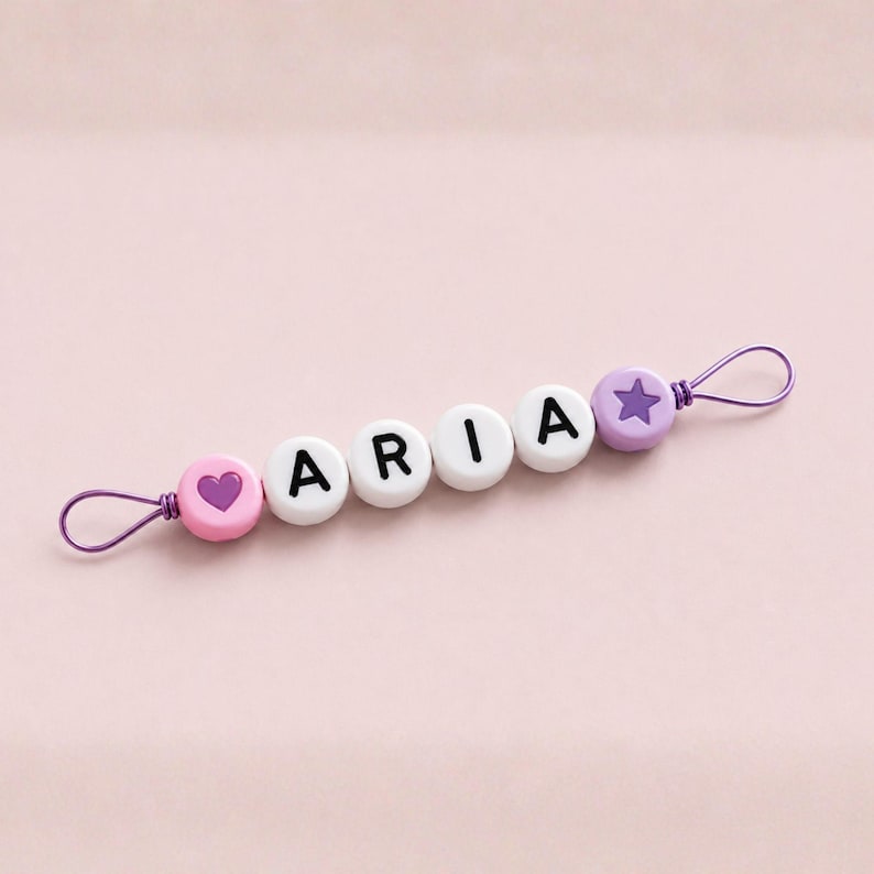 Puede incluir: Un clip para chupete de beb&eacute; con el nombre "ARIA" escrito con cuentas de letras blancas. El clip presenta una cuenta rosa con forma de coraz&oacute;n, cuentas de letras blancas y una cuenta morada con forma de estrella. Est&aacute; unido a un lazo de alambre morado.
