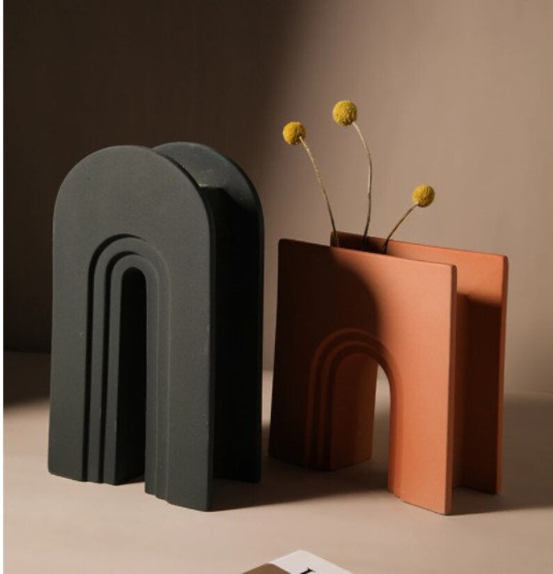 May include: Two modern ceramic vases. One is a dark gray arch shape, the other is a terracotta-colored rectangular vase with an arch cutout. The terracotta vase holds dried yellow flowers. Home decor.