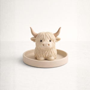 May include: A beige Highland cow figurine with a shaggy coat, curved horns, and small black eyes, sitting on a matching round tray. The figurine is a decorative item, likely for a nursery or home decor.