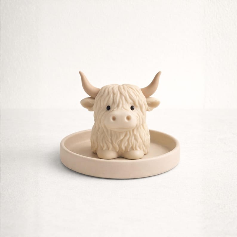 May include: A beige Highland cow figurine with a shaggy coat, curved horns, and small black eyes, sitting on a matching round tray. The figurine is a decorative item, likely for a nursery or home decor.