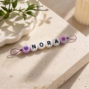 Puede incluir: Un clip de chupete personalizado con el nombre "NORA" escrito con cuentas de letras blancas. El clip presenta una cuenta morada en forma de coraz&oacute;n y estrella, y un lazo de alambre morado para sujetarlo. El fondo es una superficie texturizada neutra.