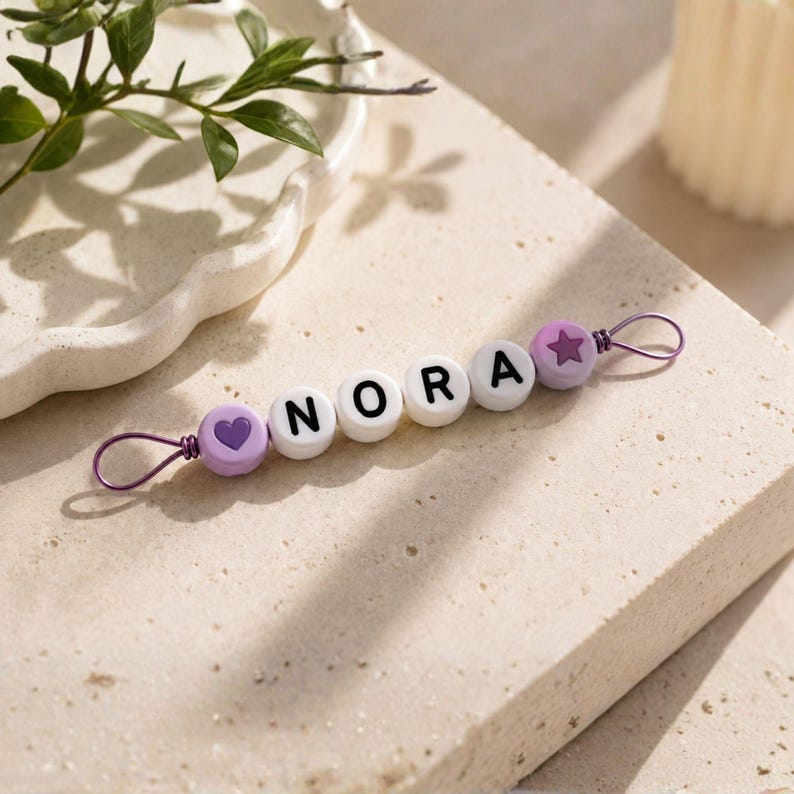 Puede incluir: Un clip de chupete personalizado con el nombre "NORA" escrito con cuentas de letras blancas. El clip presenta una cuenta morada en forma de coraz&oacute;n y estrella, y un lazo de alambre morado para sujetarlo. El fondo es una superficie texturizada neutra.
