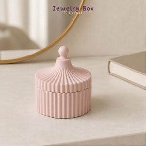 Puede incluir: Un joyero cilíndrico de color rosa claro con una tapa a juego que presenta una textura acanalada vertical y un pequeño pomo esférico en la parte superior. El texto Jewelry Box 3D STL FILES es visible en la parte superior de la imagen.