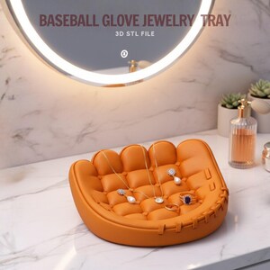 Peut inclure: Un plateau &agrave; bijoux en forme de gant de baseball orange avec un int&eacute;rieur matelass&eacute;. Le plateau contient plusieurs colliers et bagues. Le texte "BASEBALL GLOVE JEWELRY TRAY" et "3D STL FILE" est visible.