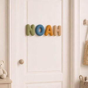 May include: A set of four individual, rounded, three-dimensional letters spelling the name NOAH, mounted on a white door. The letter N is olive green, the O is blue, the A is mustard yellow, and the H is a light beige, all featuring a smooth finish.